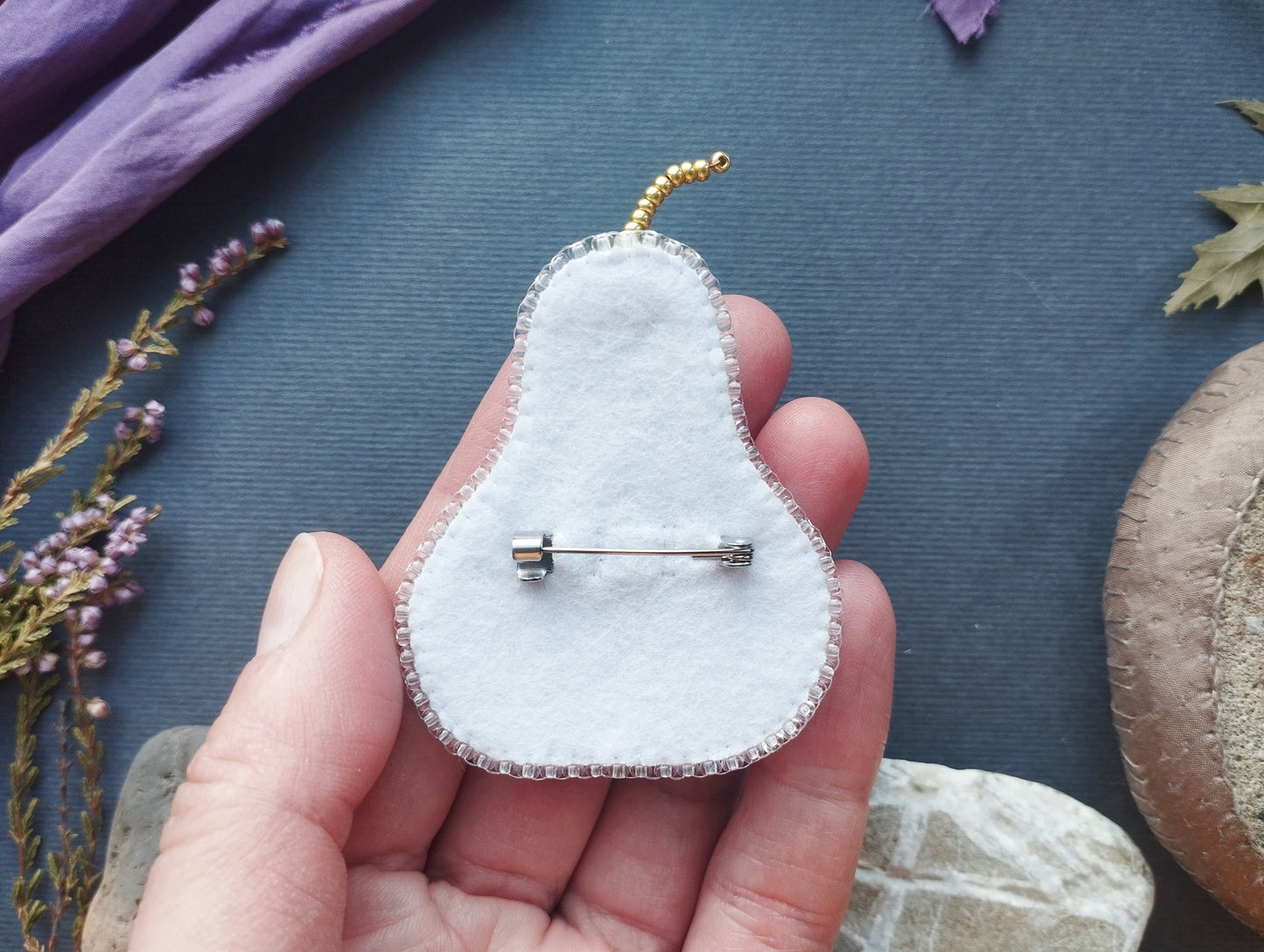 A hand holding a white felt pear pin with a silver pinback, surrounded by various objects such as flowers and a rock.