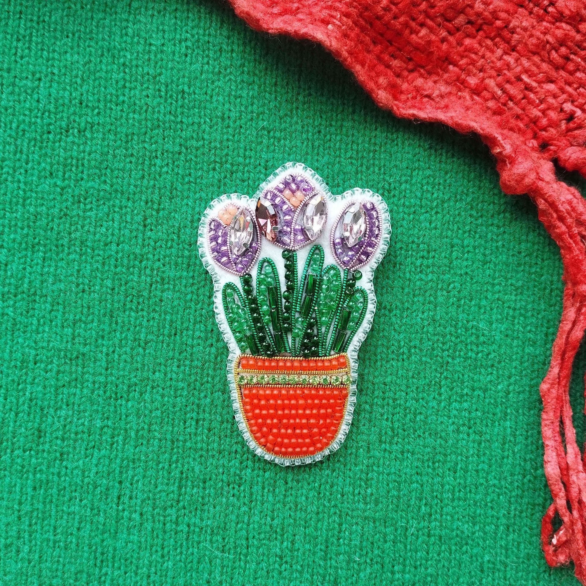A decorative pin featuring a potted plant with purple flowers and green leaves, placed on a green fabric background with a red fabric edge.