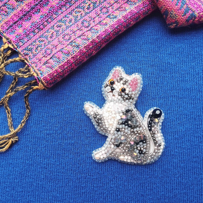 A close-up view of a beaded cat embroidered on a blue fabric surface, with a colorful woven fabric and a pink and purple patterned fabric visible in the background.