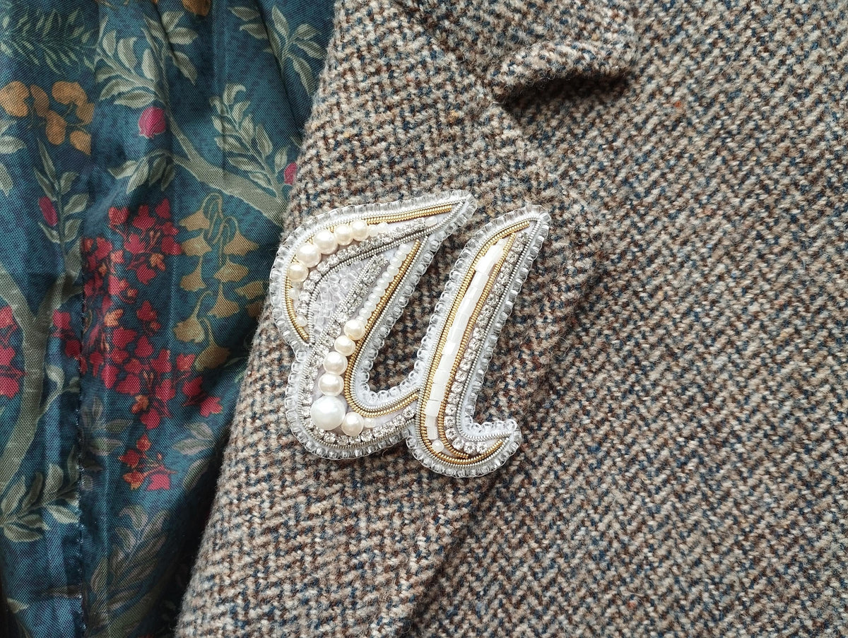 a close-up view of a decorative brooch or pin with pearls and a metallic design, attached to a tweed jacket or blazer.