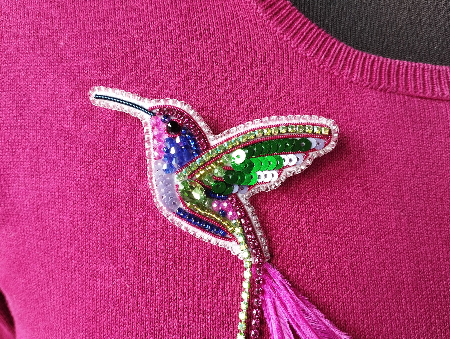 A close-up image of a pink sweater with a colorful, sequined hummingbird brooch or pin attached to it.