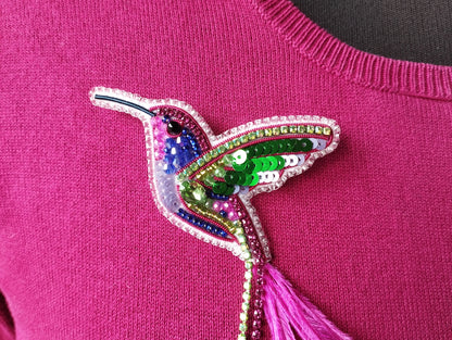 A close-up image of a pink sweater with a colorful, sequined hummingbird brooch or pin attached to it.