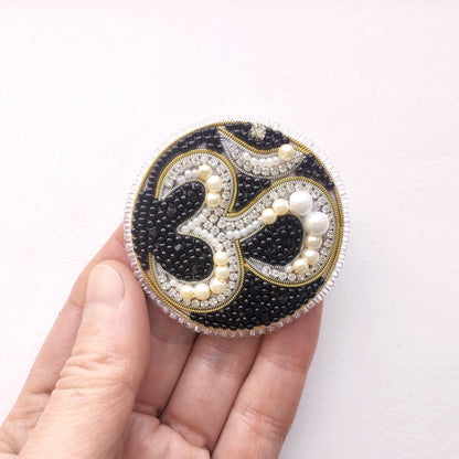 A hand holding a circular, ornate piece of jewelry with a black and white design, including pearls and beads.