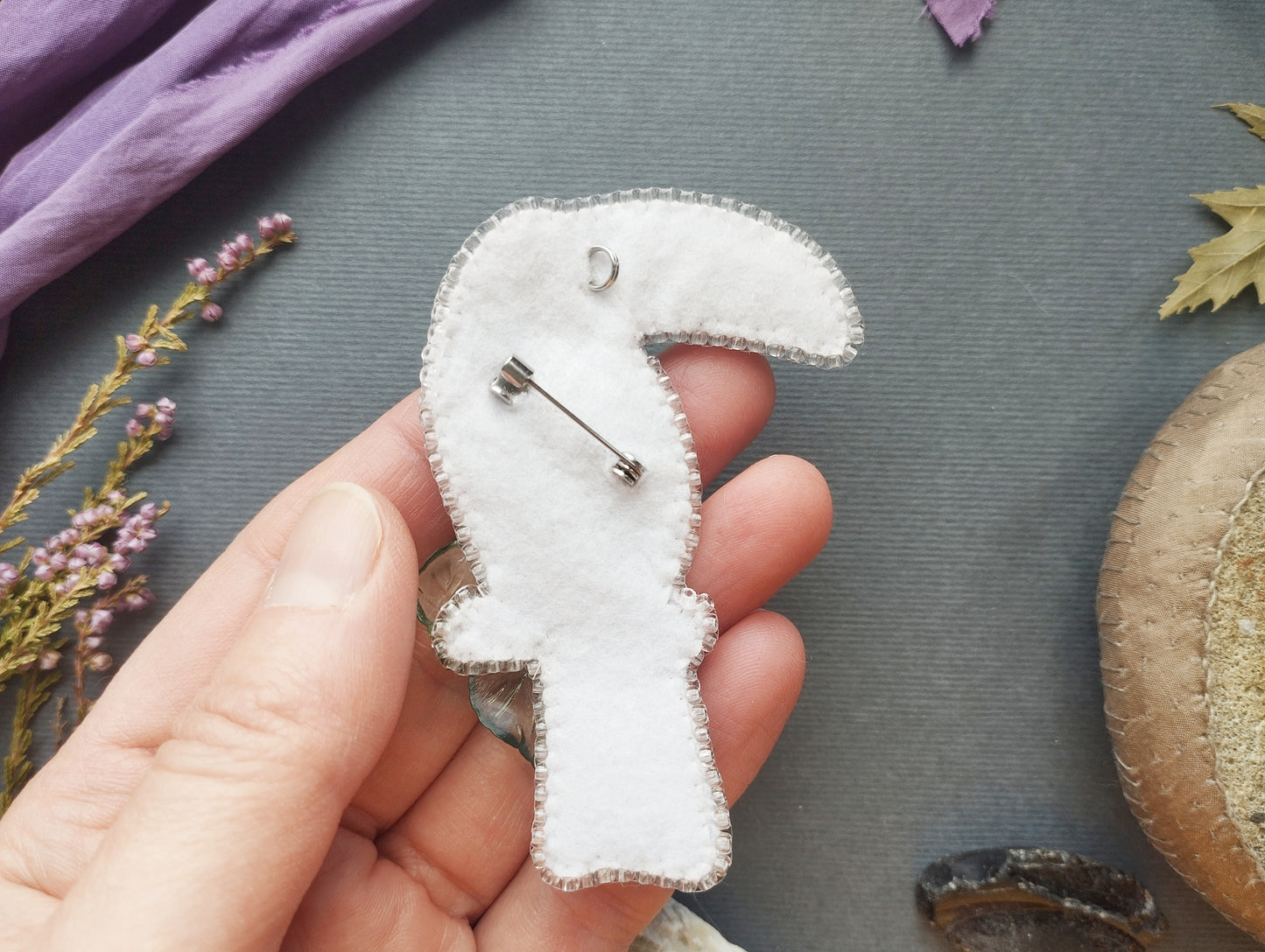 A hand holding a white felt bird pin against a gray background with various objects, including a purple fabric, flowers, and a brown object.
