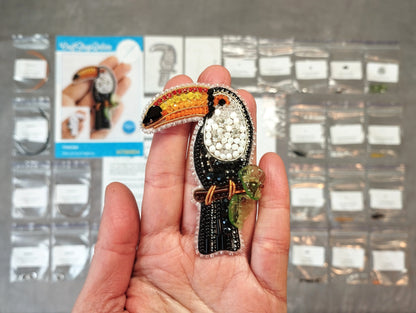 A hand holding a colorful, intricately designed bird brooch against a background of various craft supplies and materials.