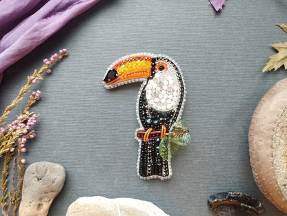 a colorful, hand-made bird brooch with a vibrant beak and wings, surrounded by various natural elements such as flowers, rocks, and leaves.
