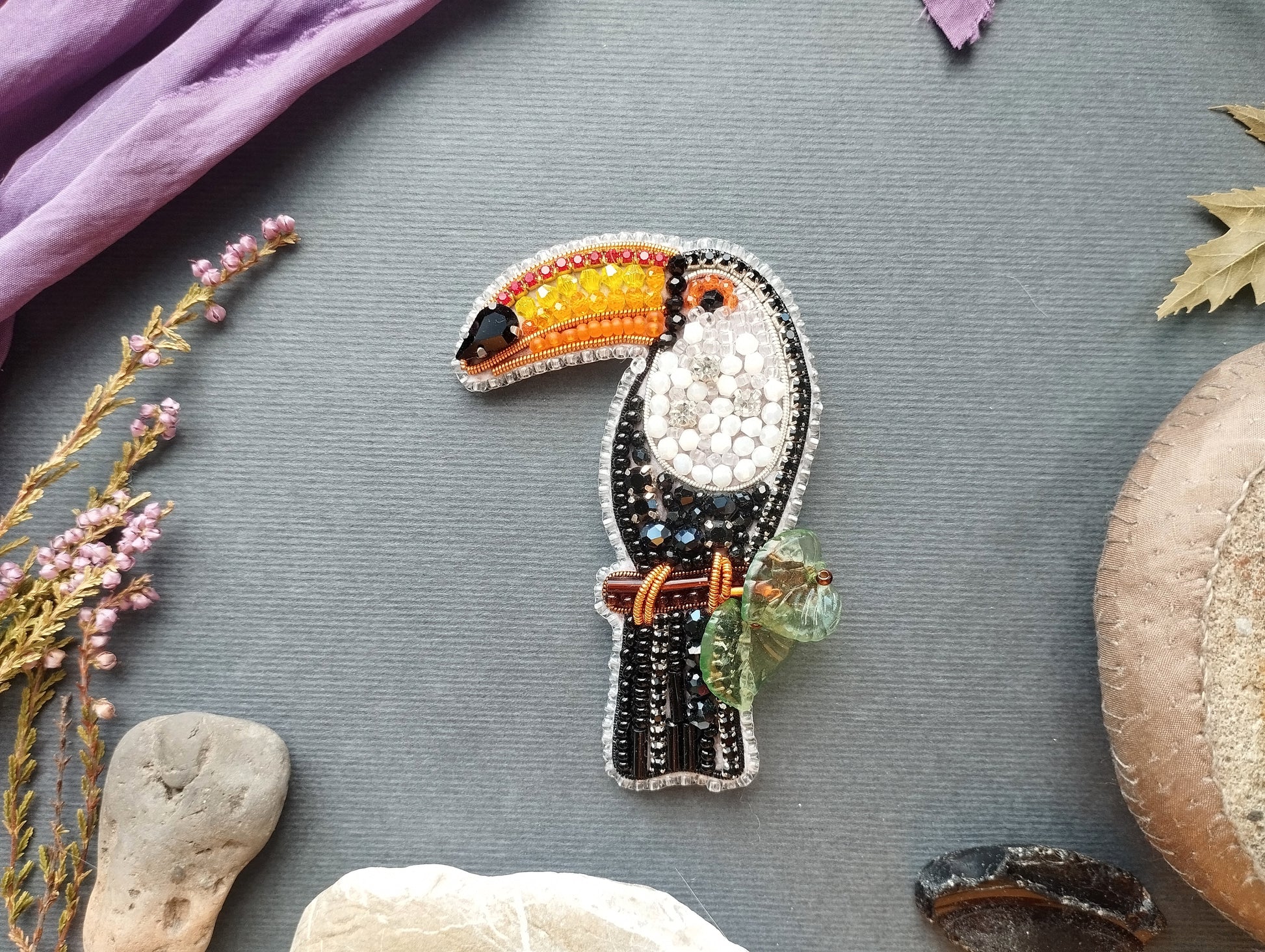 a colorful, hand-made bird brooch with a vibrant beak and wings, surrounded by various natural elements such as flowers, rocks, and leaves.