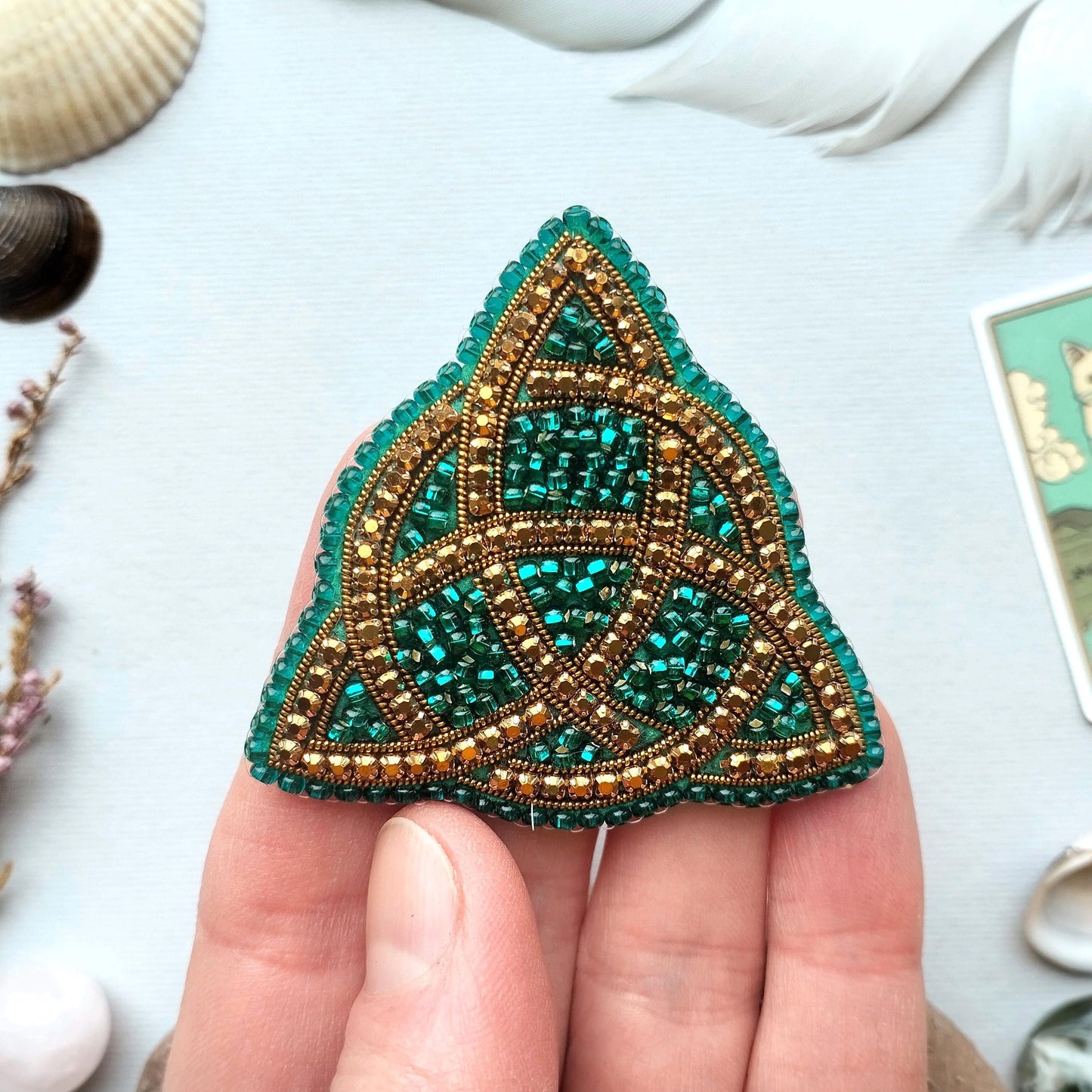 A hand holding a triangular-shaped, intricately beaded object with a green and gold color scheme.