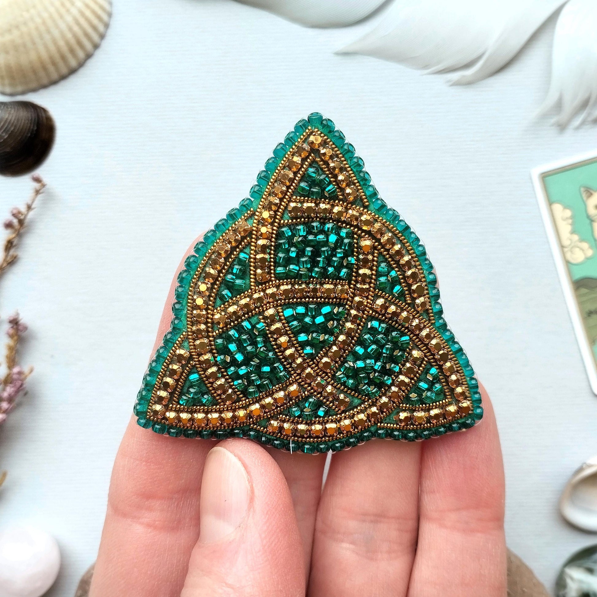 A hand holding a triangular-shaped, intricately beaded object with a green and gold color scheme.