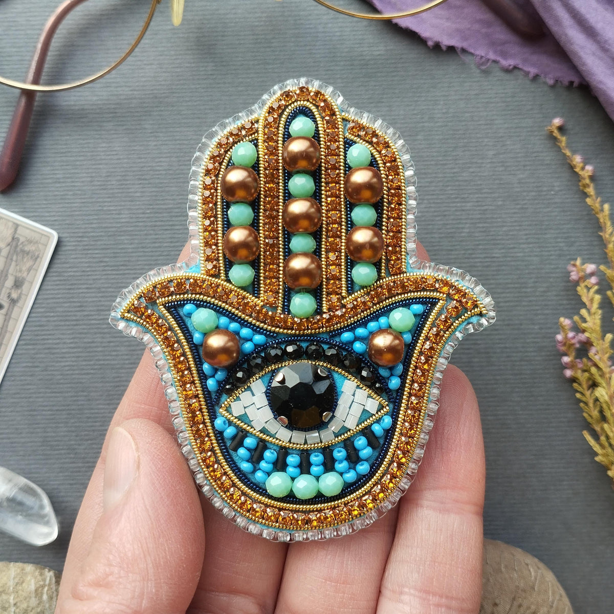 A hand holding a colorful, intricately beaded brooch or pin with a large eye design at the center.