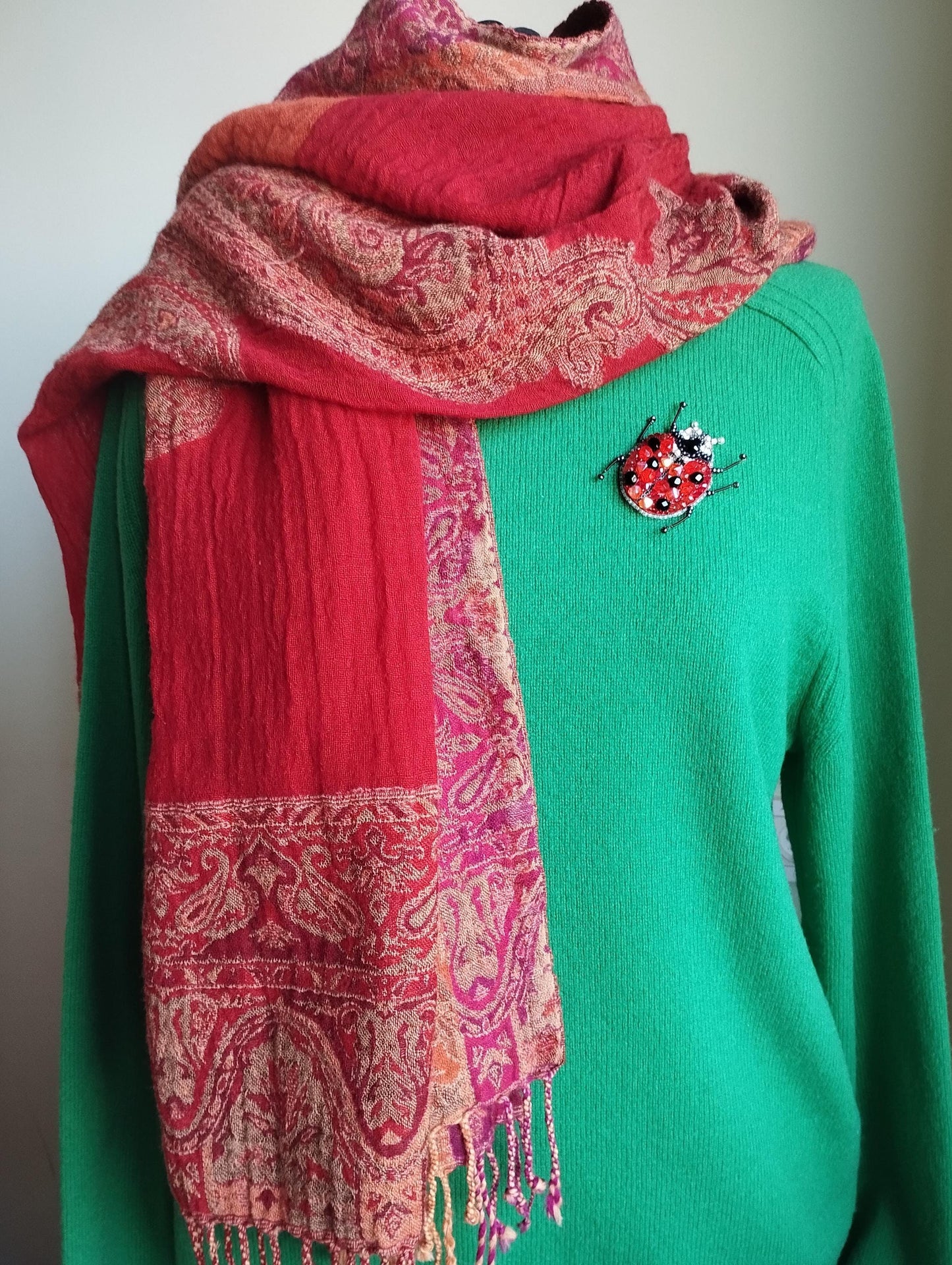 A person wearing a green sweater with a red and pink patterned scarf draped over it, and a small red and gold brooch on the sweater.
