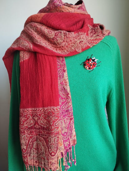 A person wearing a green sweater with a red and pink patterned scarf draped over it, and a small red and gold brooch on the sweater.
