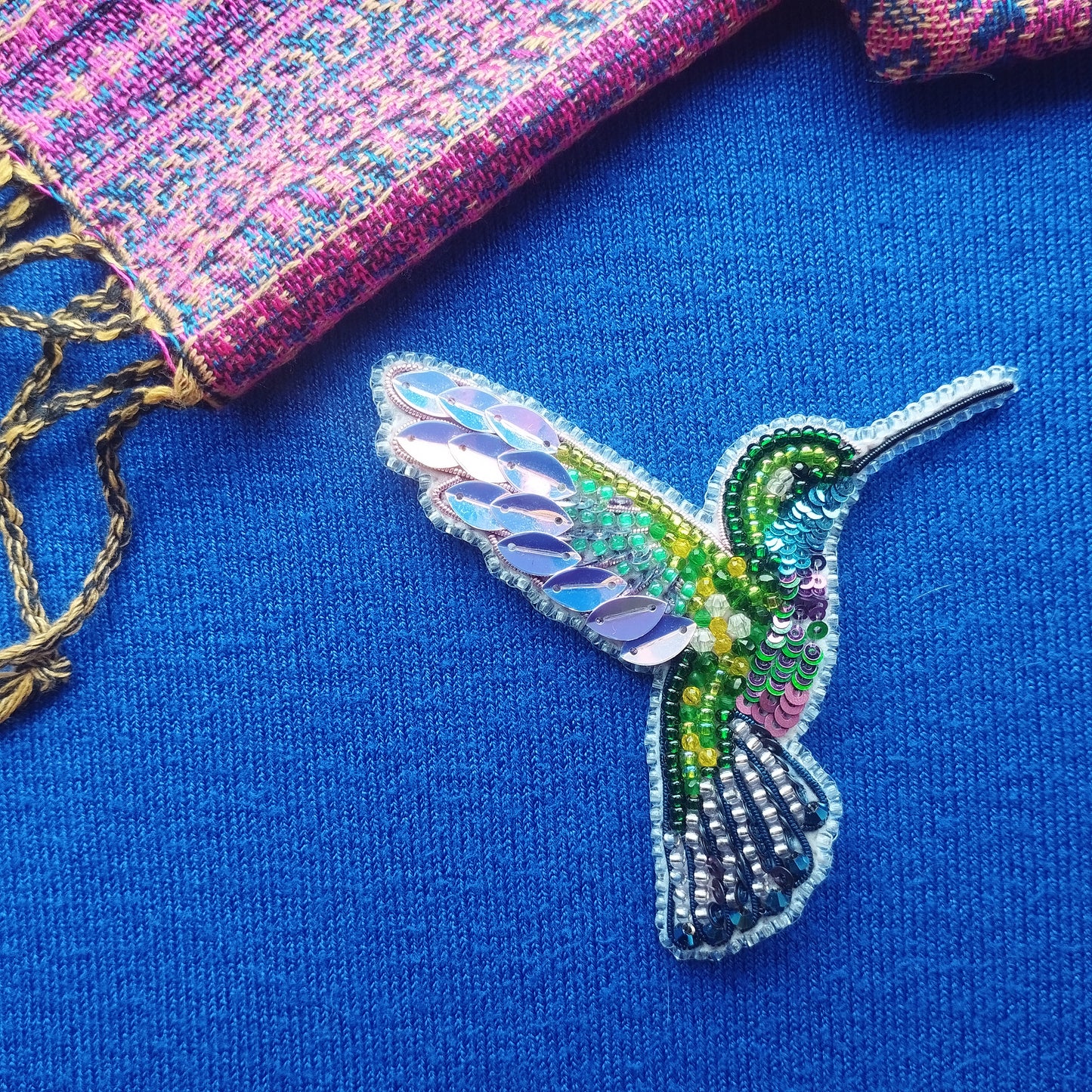 A colorful, intricately designed hummingbird brooch made of beads and sequins is displayed against a blue background.
