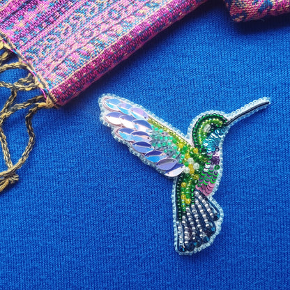 A colorful, intricately designed hummingbird brooch made of beads and sequins is displayed against a blue background.