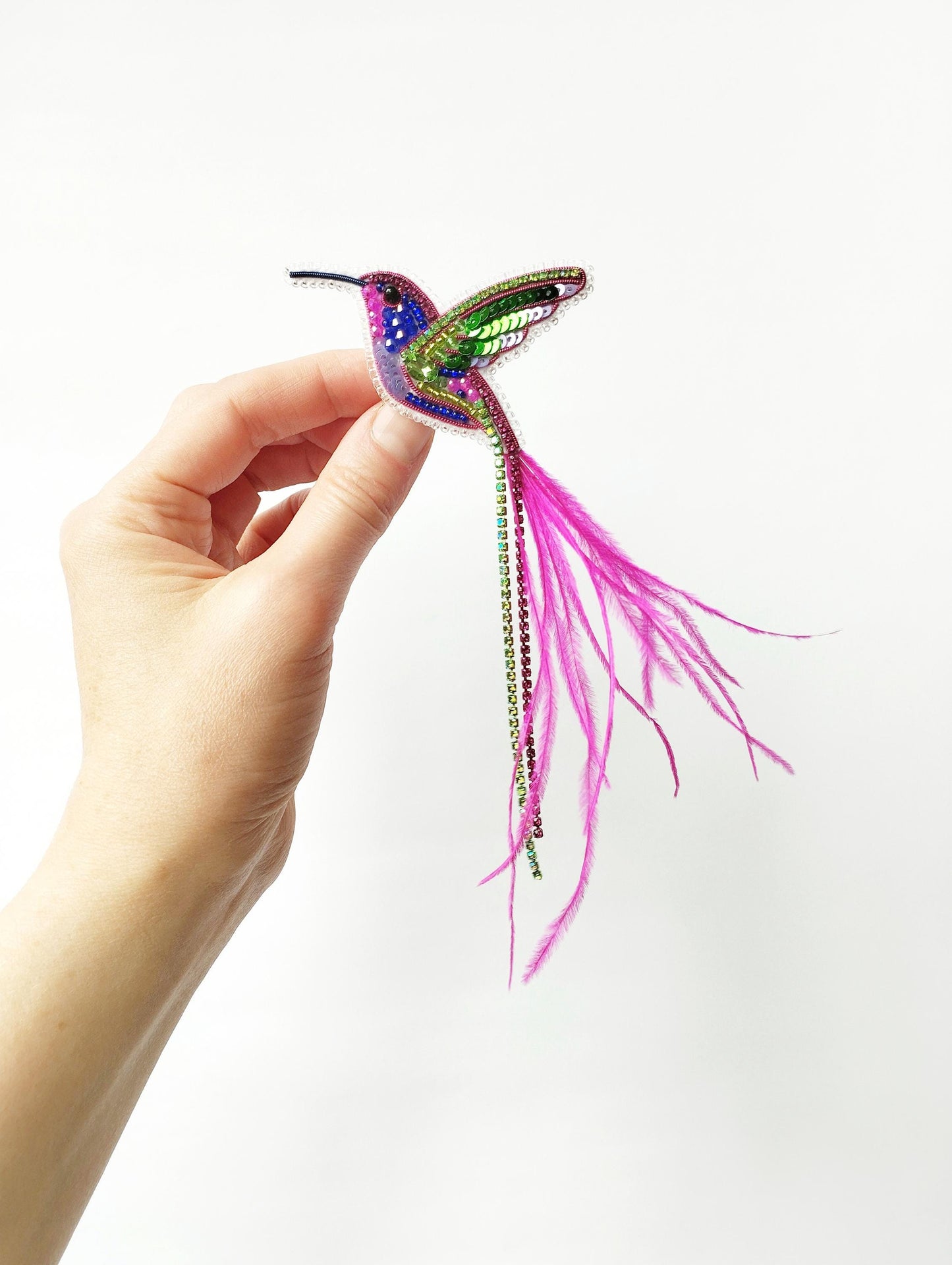A hand is holding a colorful, intricately designed hummingbird brooch against a white background.