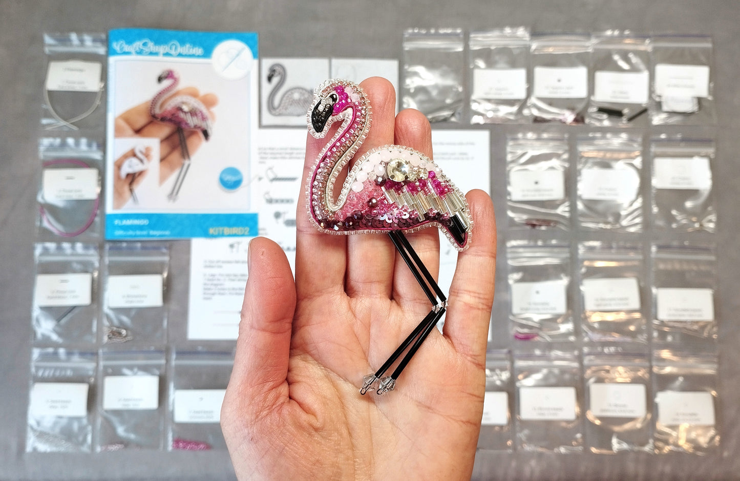 A hand holding a small, intricately beaded flamingo brooch against a background of various sewing supplies and materials.