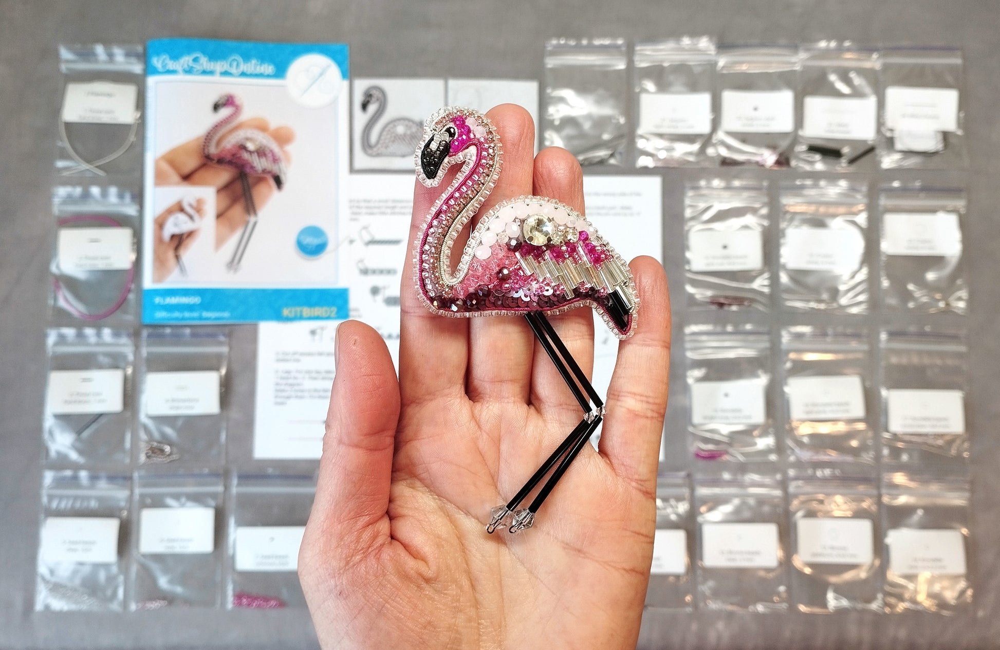 A hand holding a small, intricately beaded flamingo brooch against a background of various sewing supplies and materials.