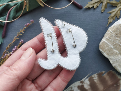 A hand holding a white fabric pin with silver buttons, surrounded by various objects such as a pair of glasses, leaves, and feathers.