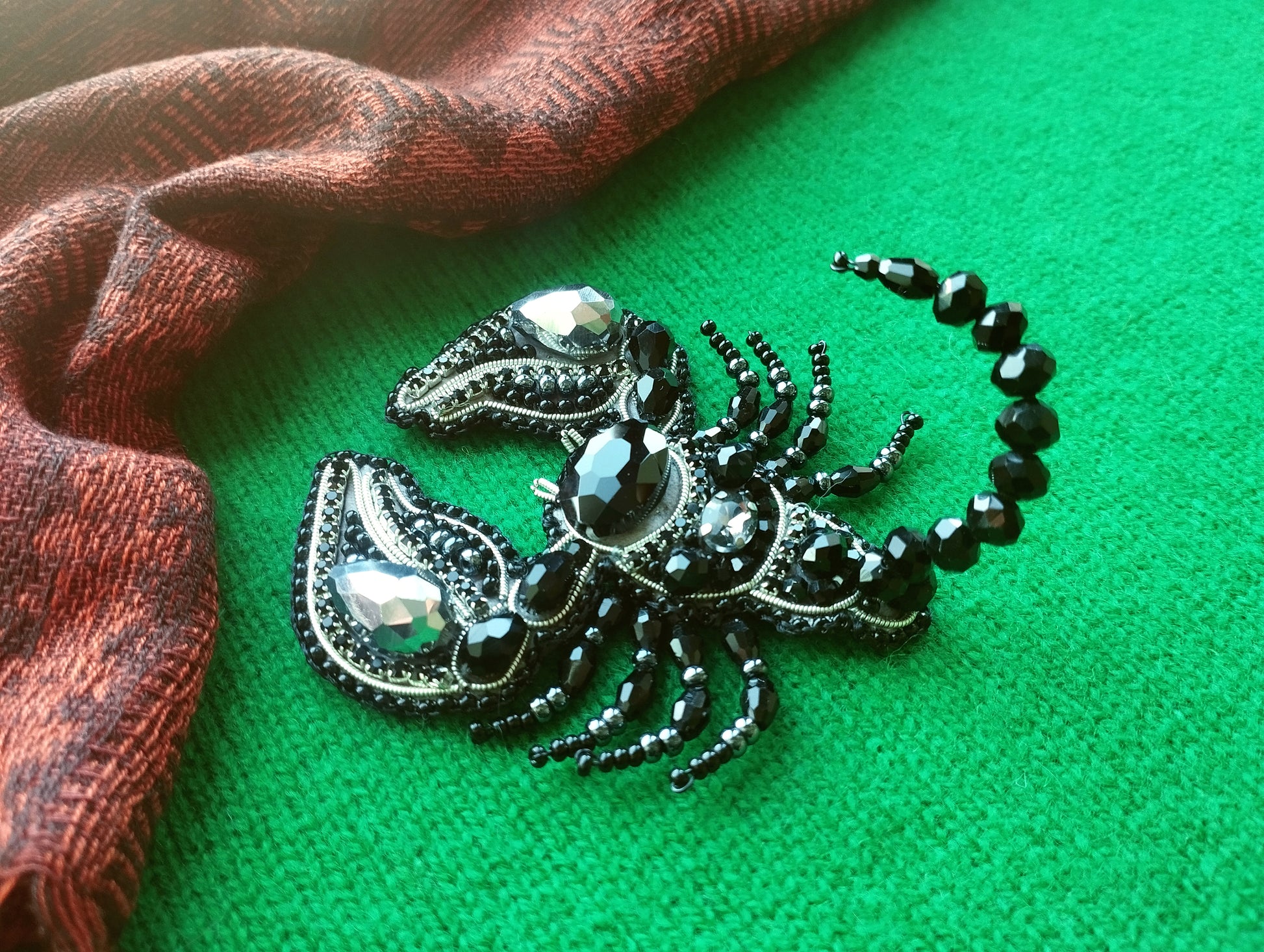 A decorative black and silver piece of jewelry, possibly a brooch or pendant, is displayed on a green surface next to a red and black patterned fabric.