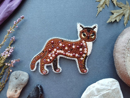 a detailed embroidered or beaded cat design on a blue background, surrounded by various natural elements such as leaves, rocks, and flowers.