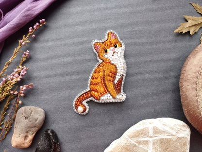 a decorative embroidered cat piece placed on a grey background, surrounded by various natural elements such as leaves, flowers, and rocks.