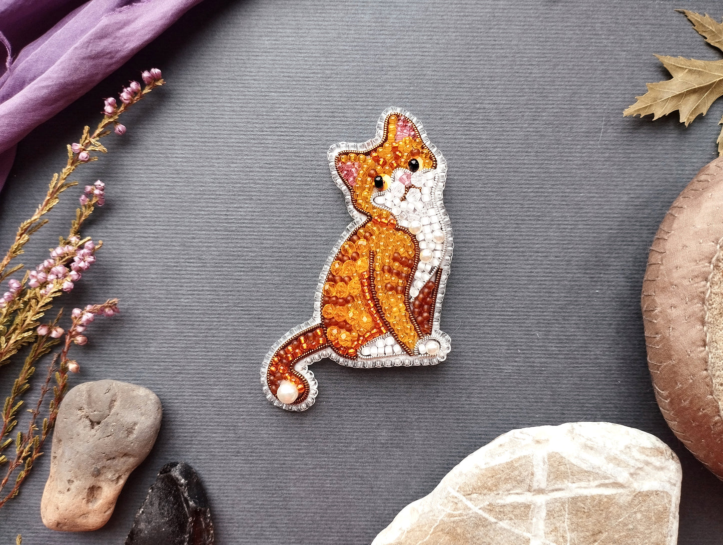 a decorative embroidered cat piece placed on a grey background, surrounded by various natural elements such as leaves, flowers, and rocks.