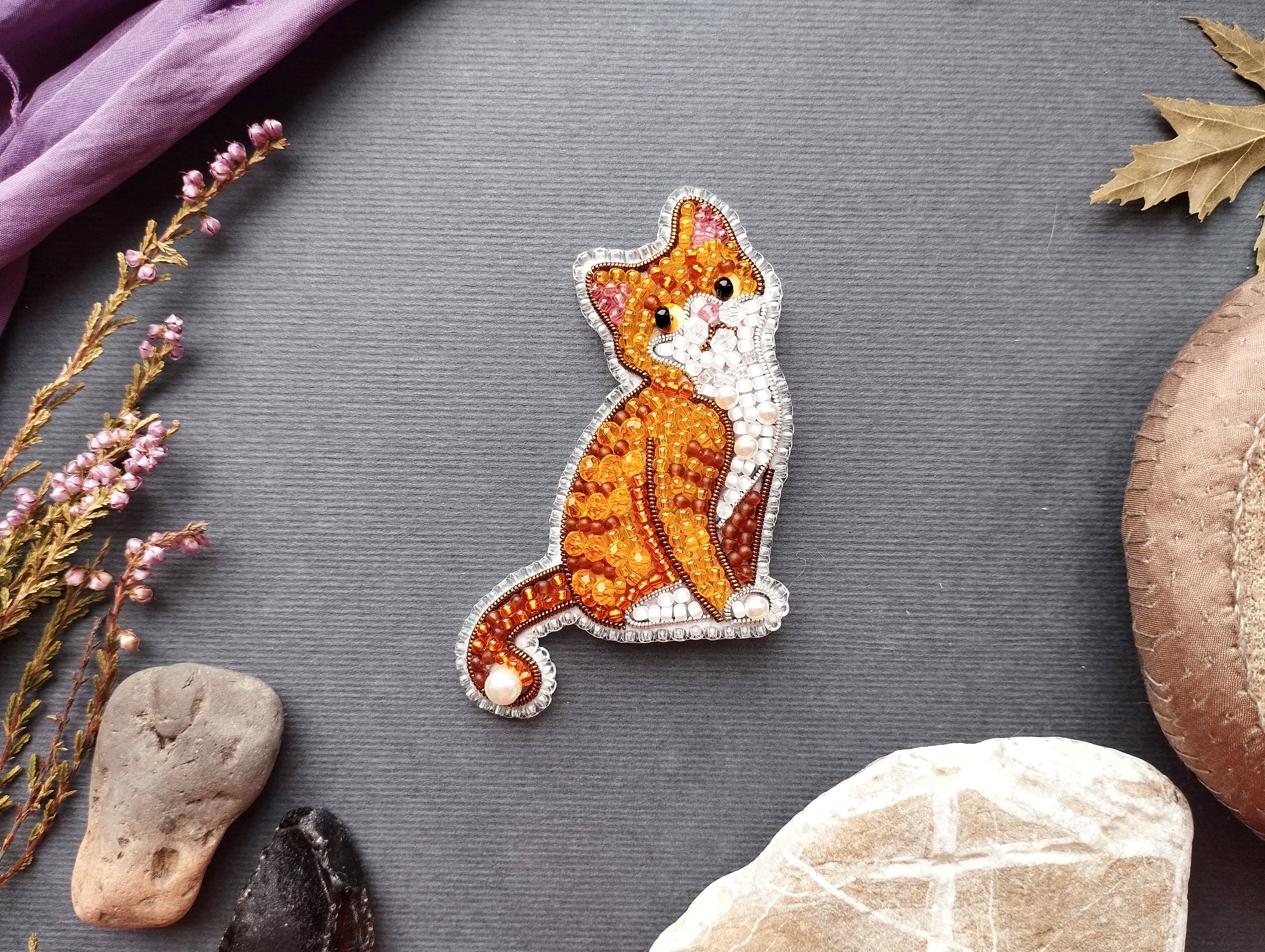 a decorative embroidered cat piece placed on a grey background, surrounded by various natural elements such as leaves, flowers, and rocks.