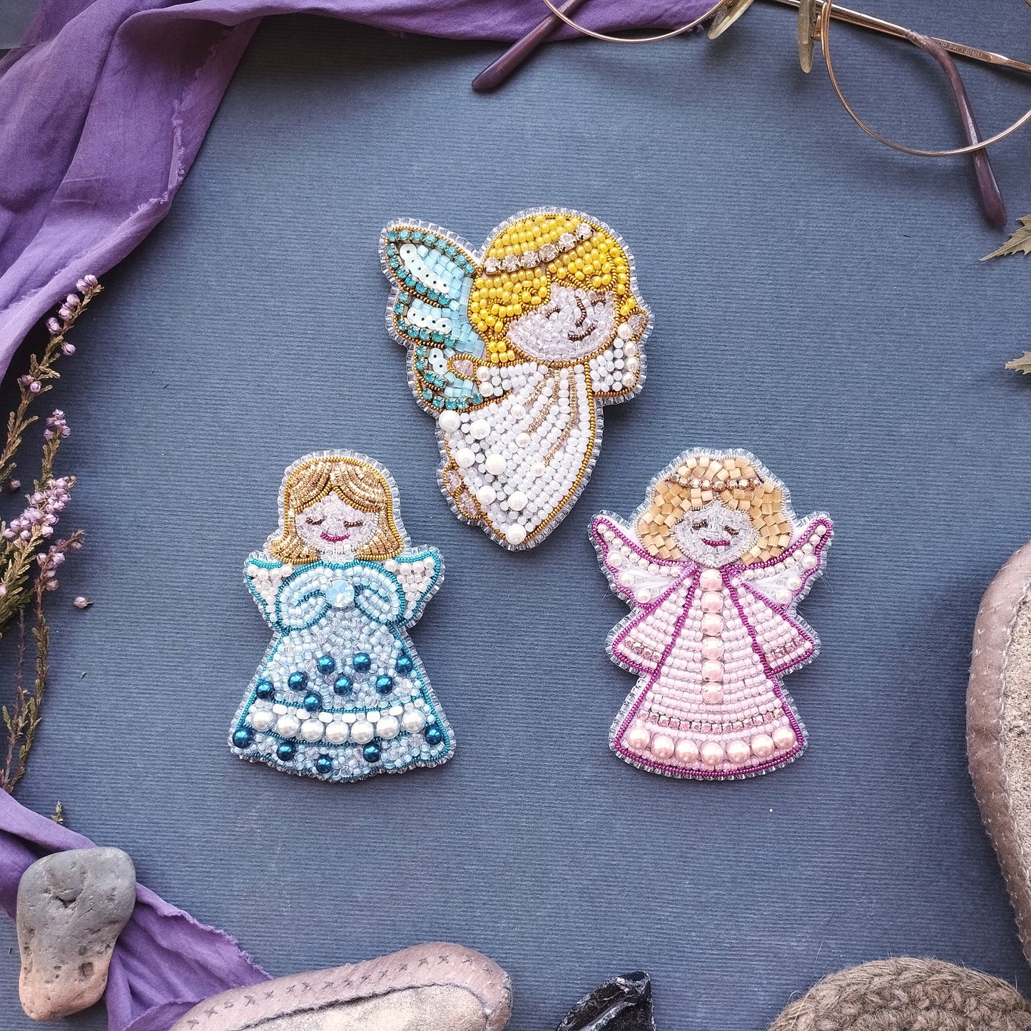 three beaded angel brooches with different colored dresses, arranged on a blue background.