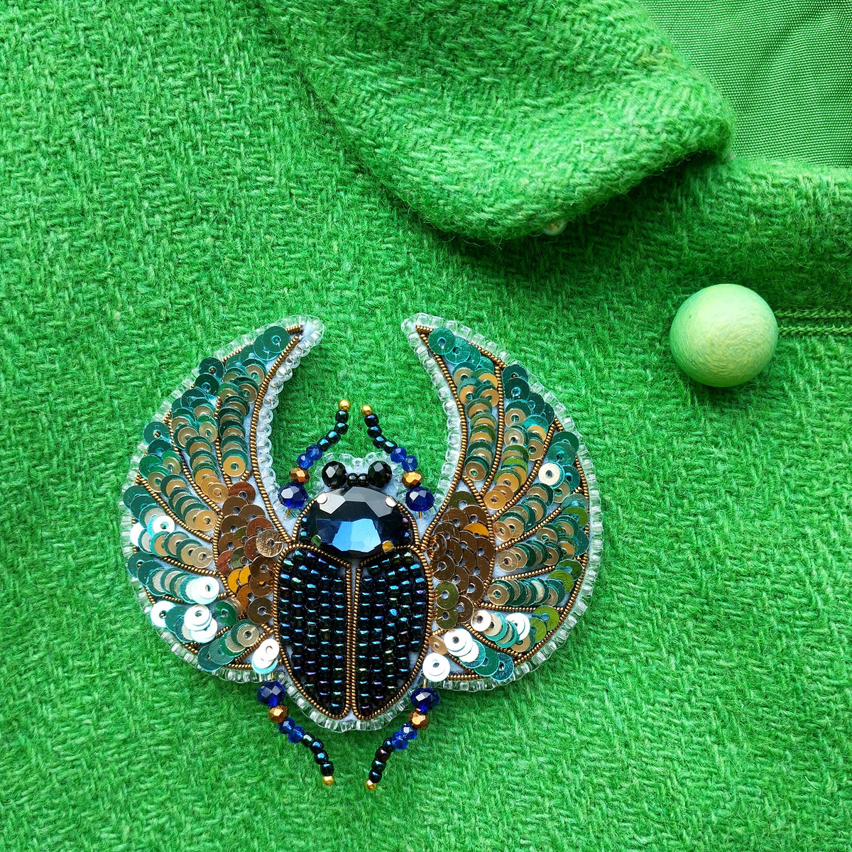 a close-up view of a decorative piece, possibly a brooch or pin, with a vibrant green background. The piece appears to be made of beads and sequins, creating a visually striking contrast against the green fabric.