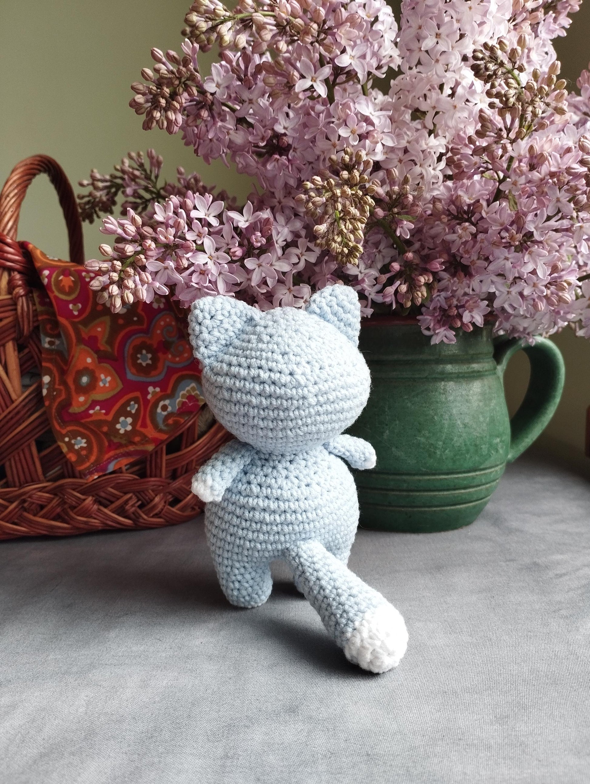 a crocheted teddy bear sitting next to a basket of lilacs