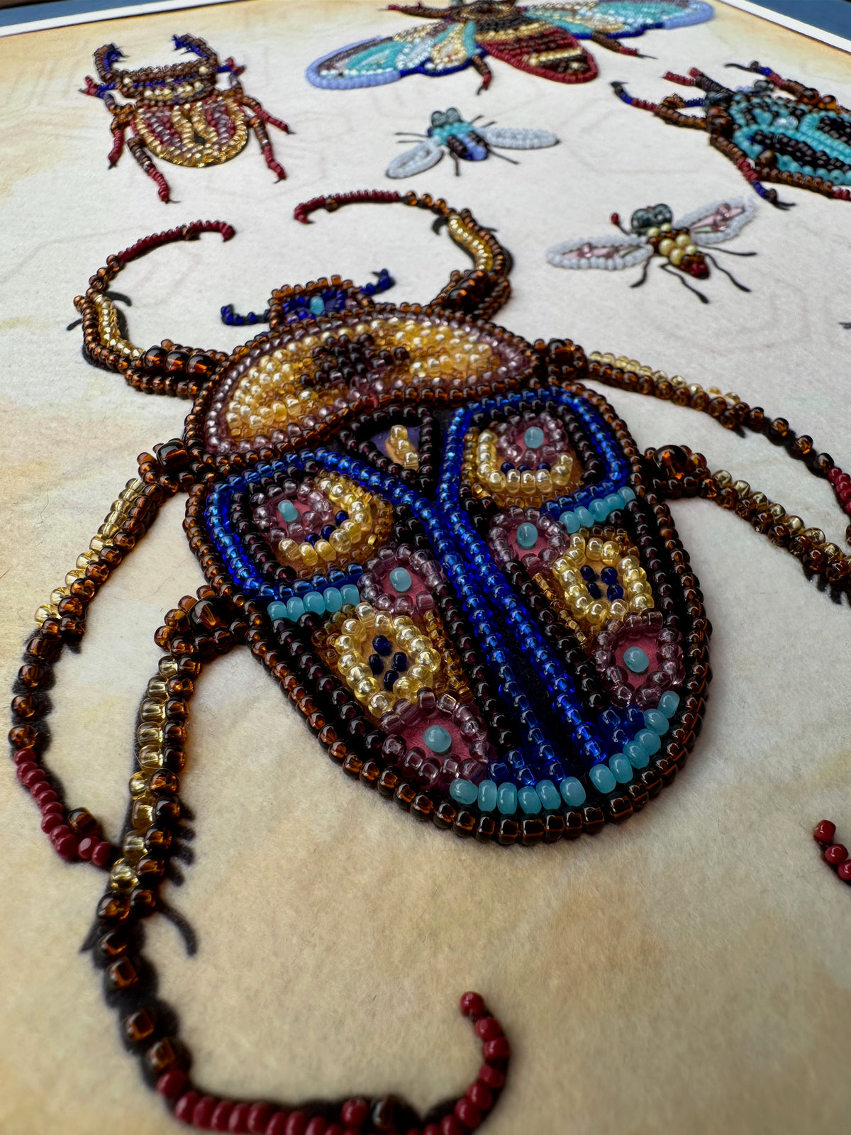 a close-up view of a detailed embroidered design of a large, colorful beetle. The design is composed of numerous small beads in various colors, creating a vibrant and intricate pattern. The image showcases the craftsmanship and artistry involved in creating such a detailed and visually appealing piece of embroidery.