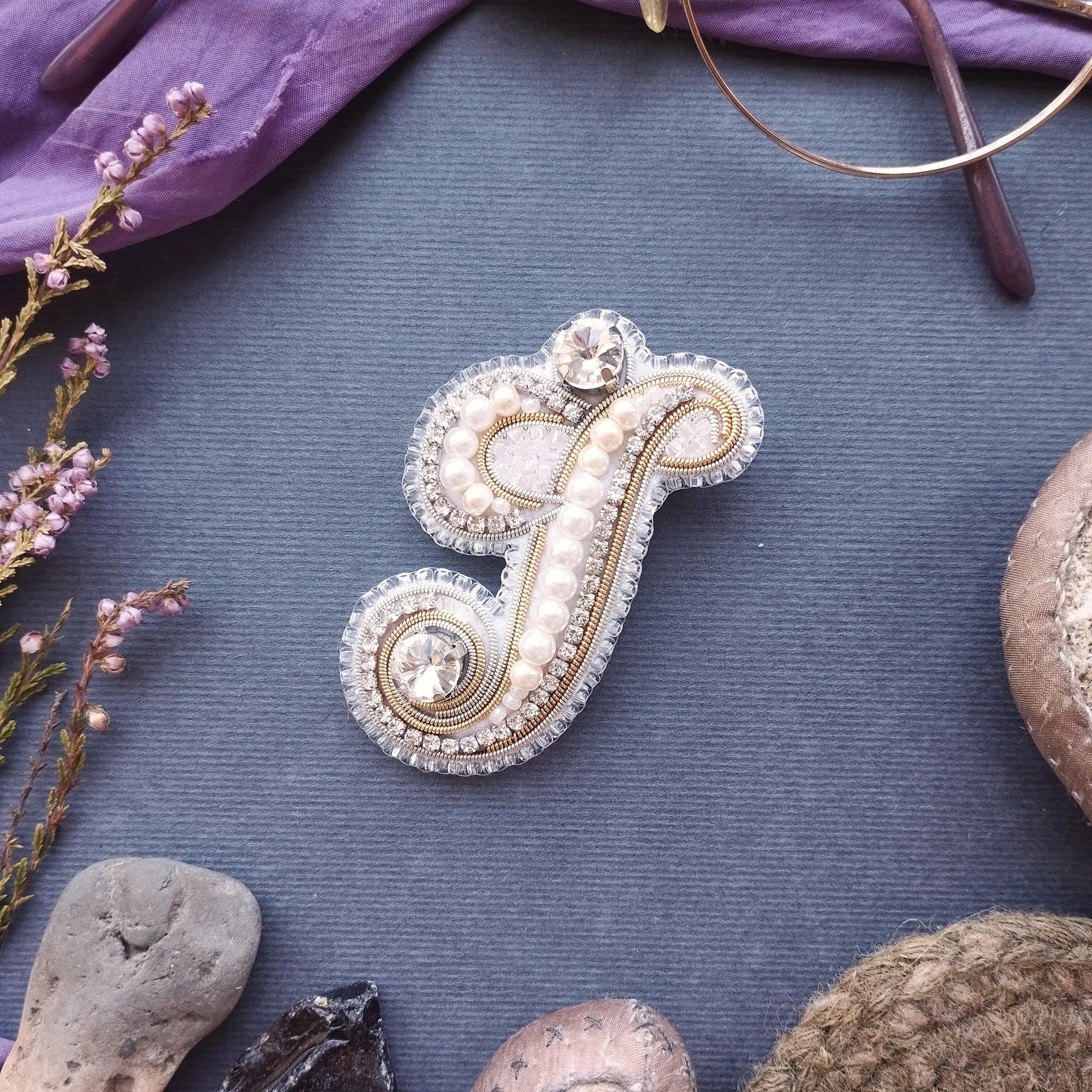 a decorative monogrammed letter 'F' adorned with pearls and rhinestones, surrounded by various natural elements such as flowers, rocks, and a pair of glasses.