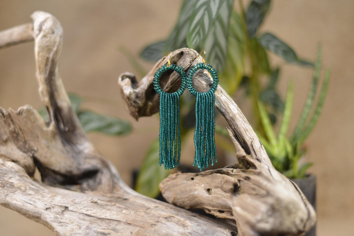 A pair of green earrings with tassels hanging from a wooden branch, with a plant in the background.
