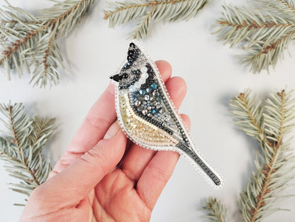 a hand holding a bird brooch with a pine branch in the background