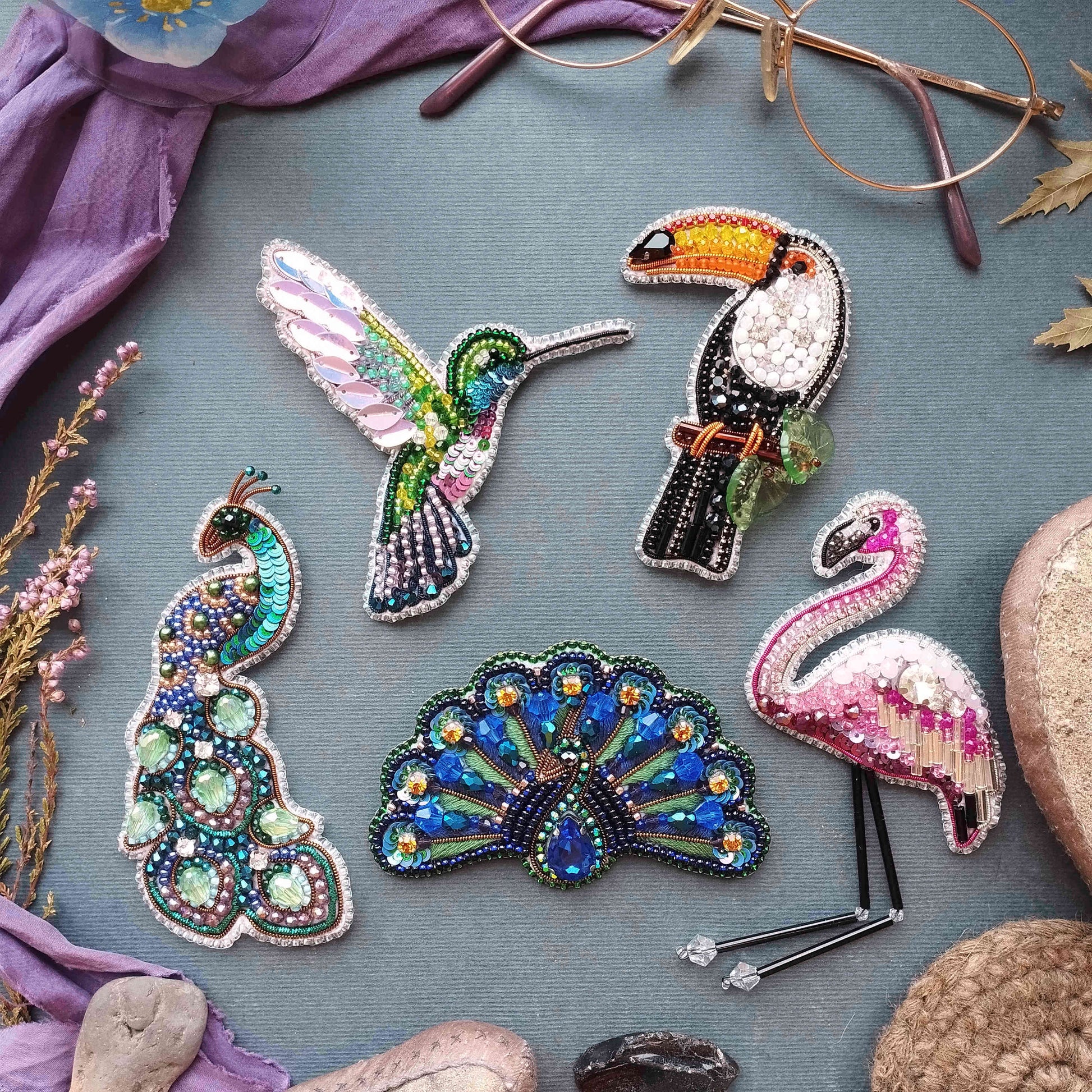 a collection of colorful, intricately designed beaded or embroidered animal pins or brooches, including a hummingbird, a peacock, a flamingo, and a parrot, all set against a purple background.