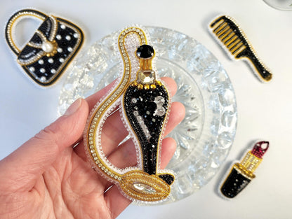 A hand holding a decorative piece of jewelry, which appears to be a perfume bottle or perfume bottle-shaped object, surrounded by other decorative items such as a purse and a brush.