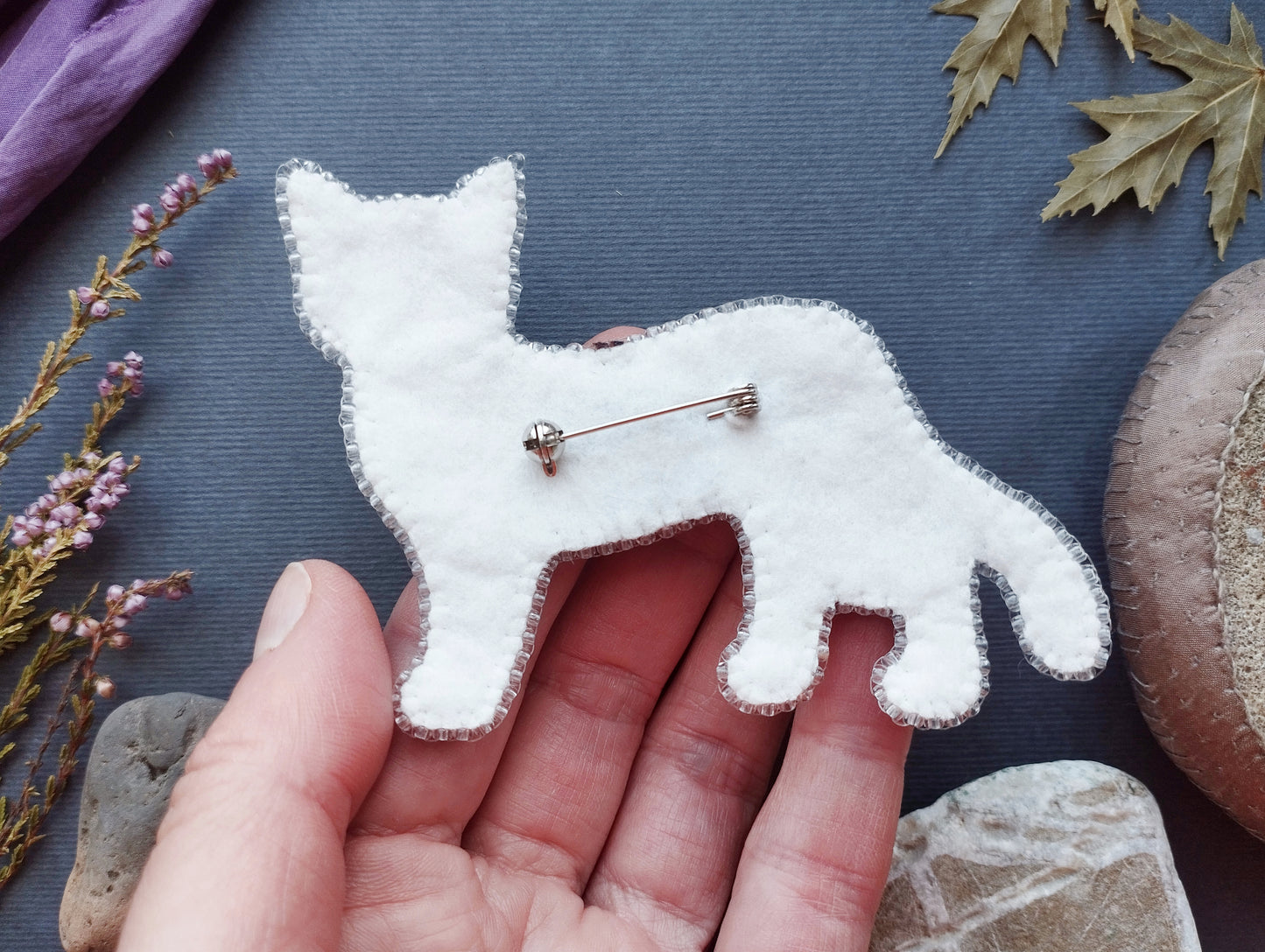 A hand holding a white felt cat pin against a dark background with various natural elements, such as leaves and rocks.
