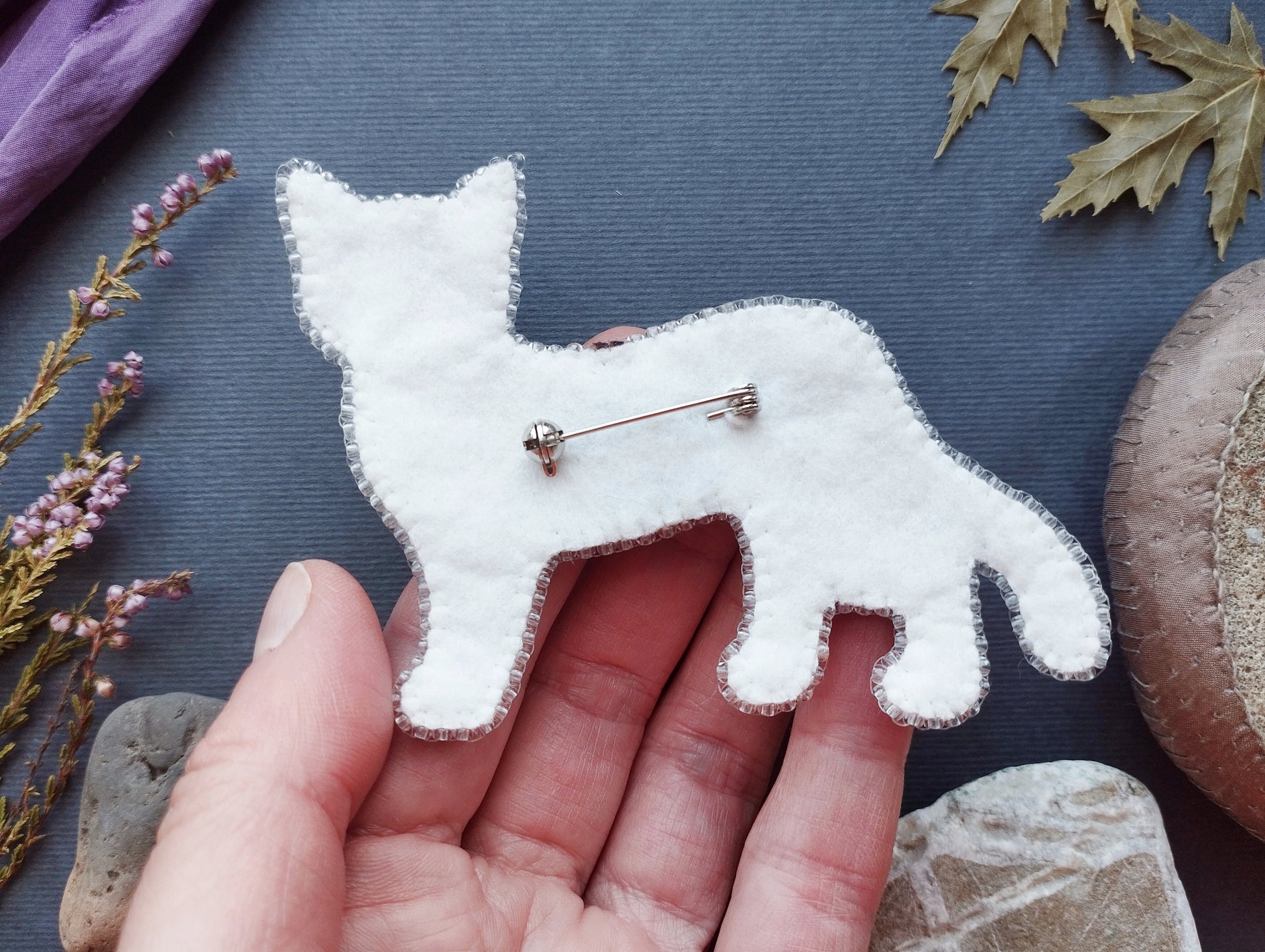 A hand holding a white felt cat pin against a dark background with various natural elements, such as leaves and rocks.