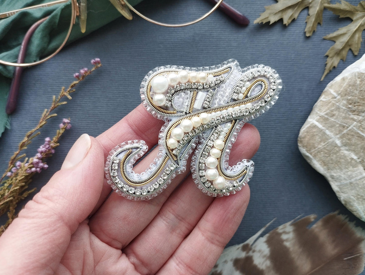 A hand holding a decorative letter brooch made of silver, white, and gold beads, with a floral design.