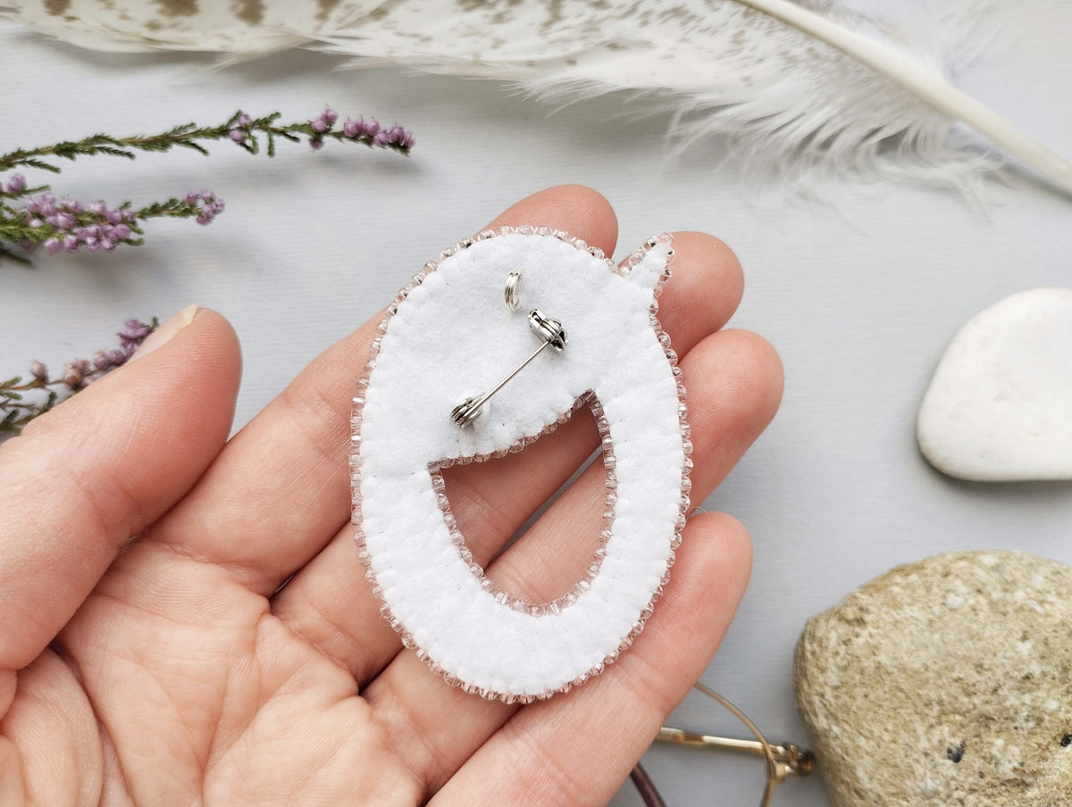 A hand holding a white fabric piece with a silver pin attached to it, surrounded by various natural elements such as feathers, flowers, and rocks.