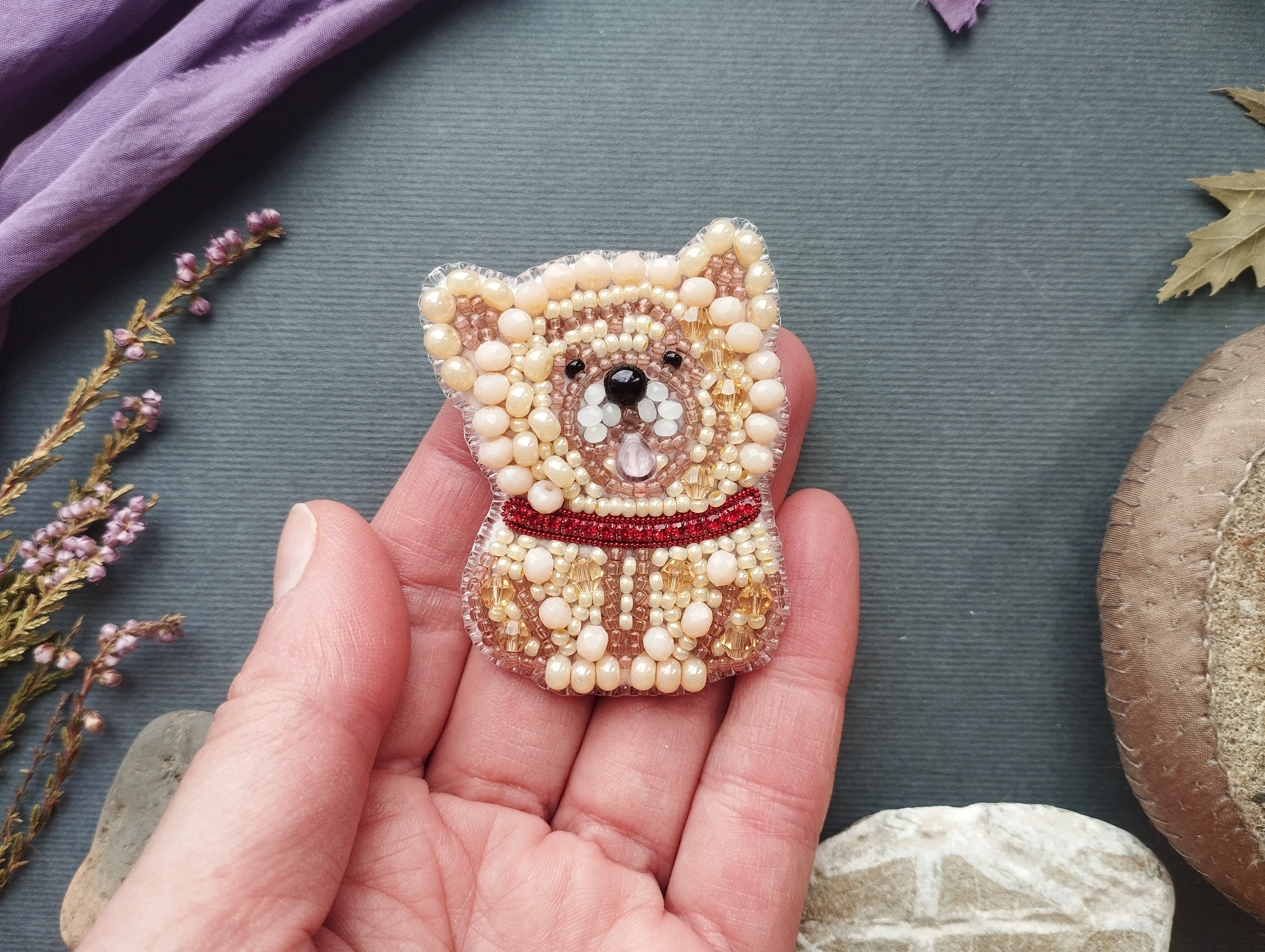 A hand holding a small, intricately beaded pin or brooch in the shape of a dog, with a red collar. The background includes various objects such as flowers, rocks, and a purple fabric.