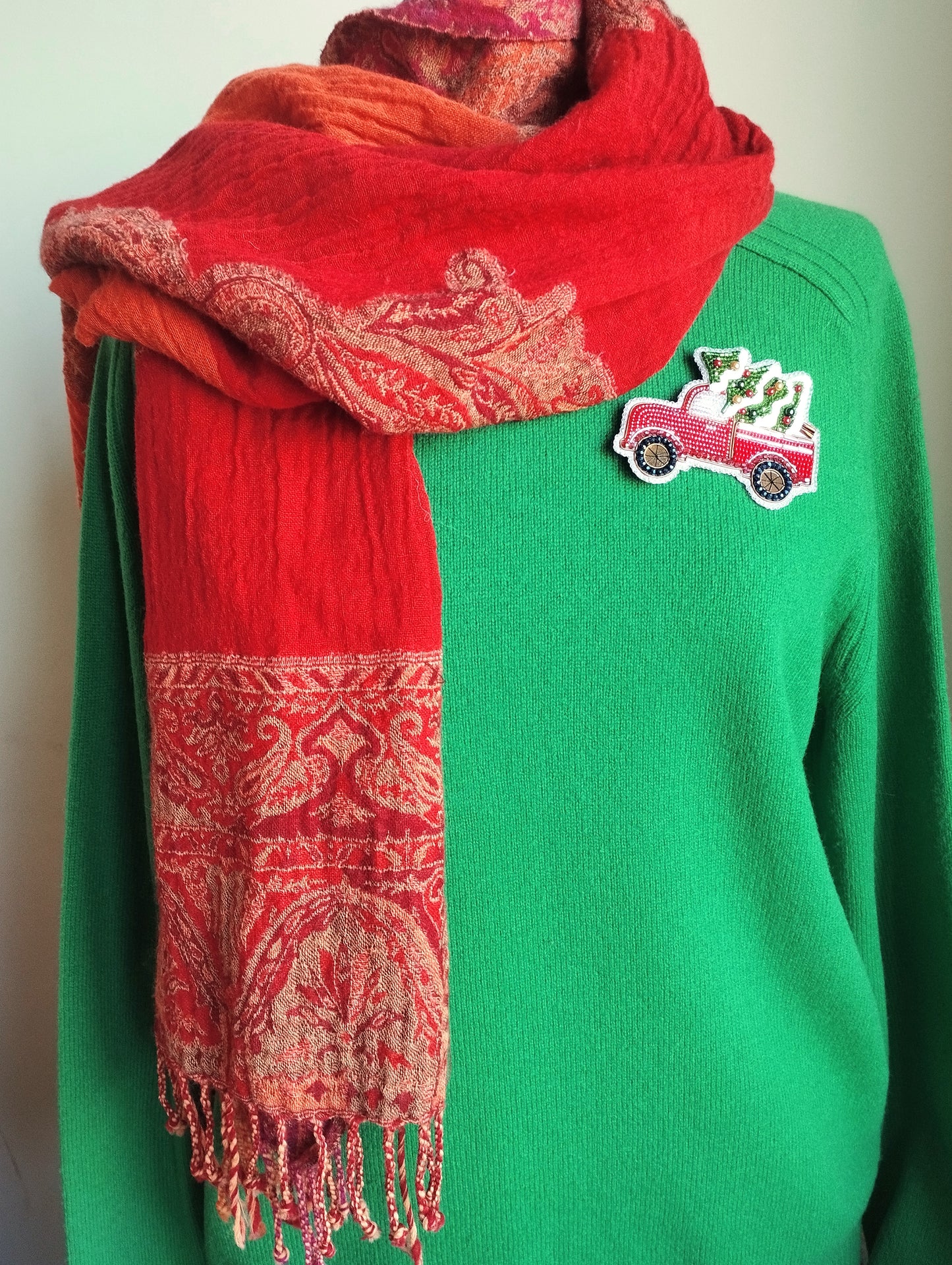 A person wearing a green sweater with a red and orange patterned scarf draped over it, and a small patch on the sweater featuring a red truck.