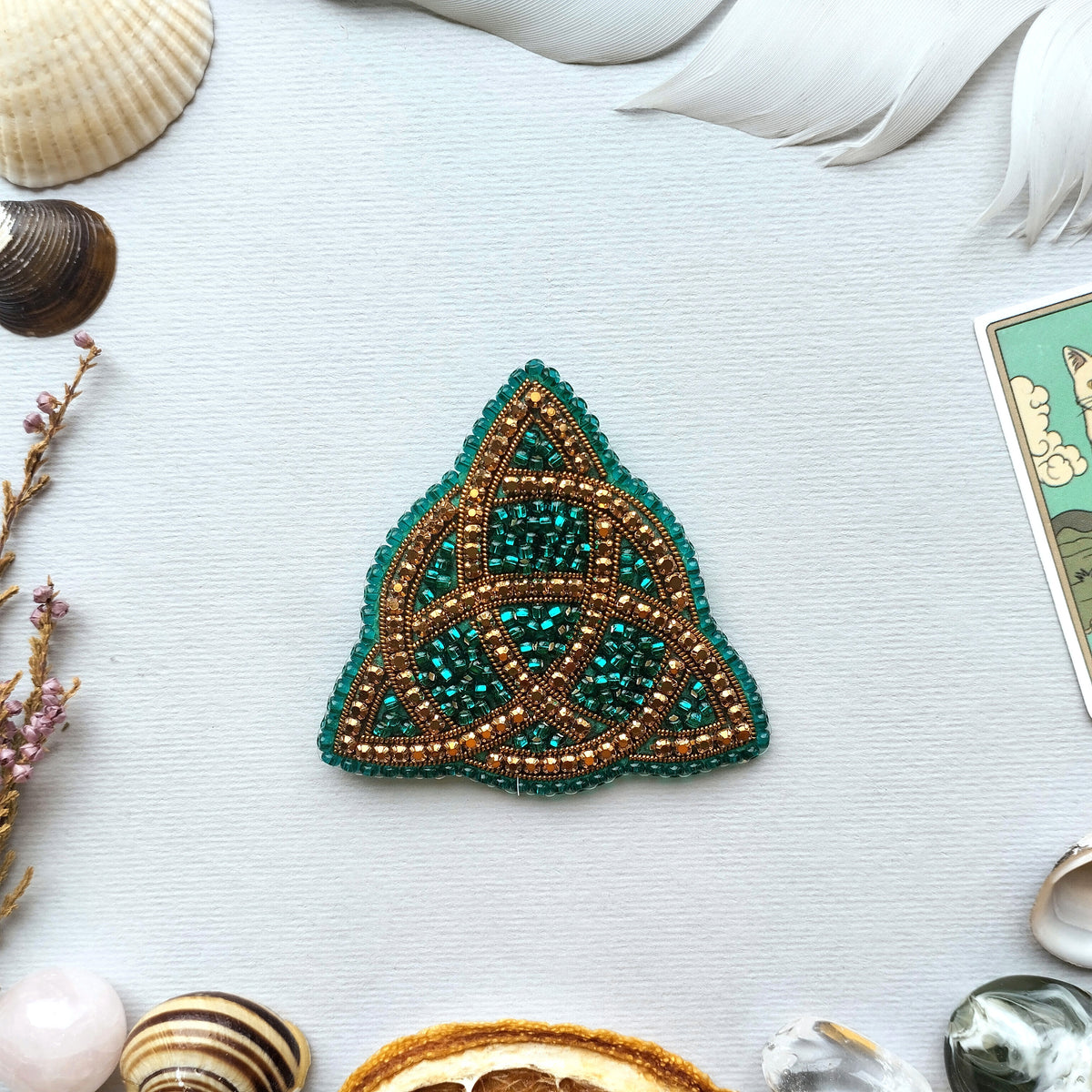 a triangular piece of jewelry with a green and gold design, surrounded by various natural elements such as seashells, feathers, and a card.