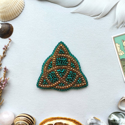a triangular piece of jewelry with a green and gold design, surrounded by various natural elements such as seashells, feathers, and a card.