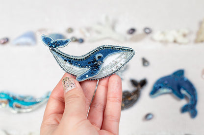 A hand holding a decorative blue and white beaded object resembling a whale or dolphin, with other similar objects in the background.