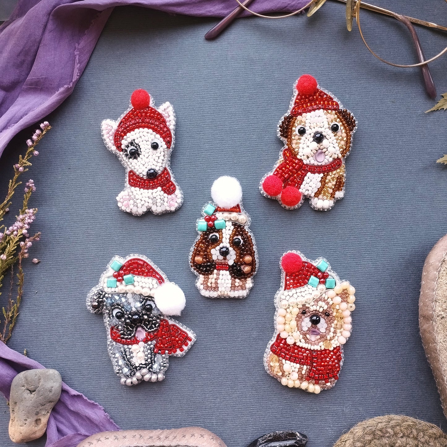 five small, intricately designed dog brooches or pins, each wearing a red hat and scarf, arranged on a gray background.