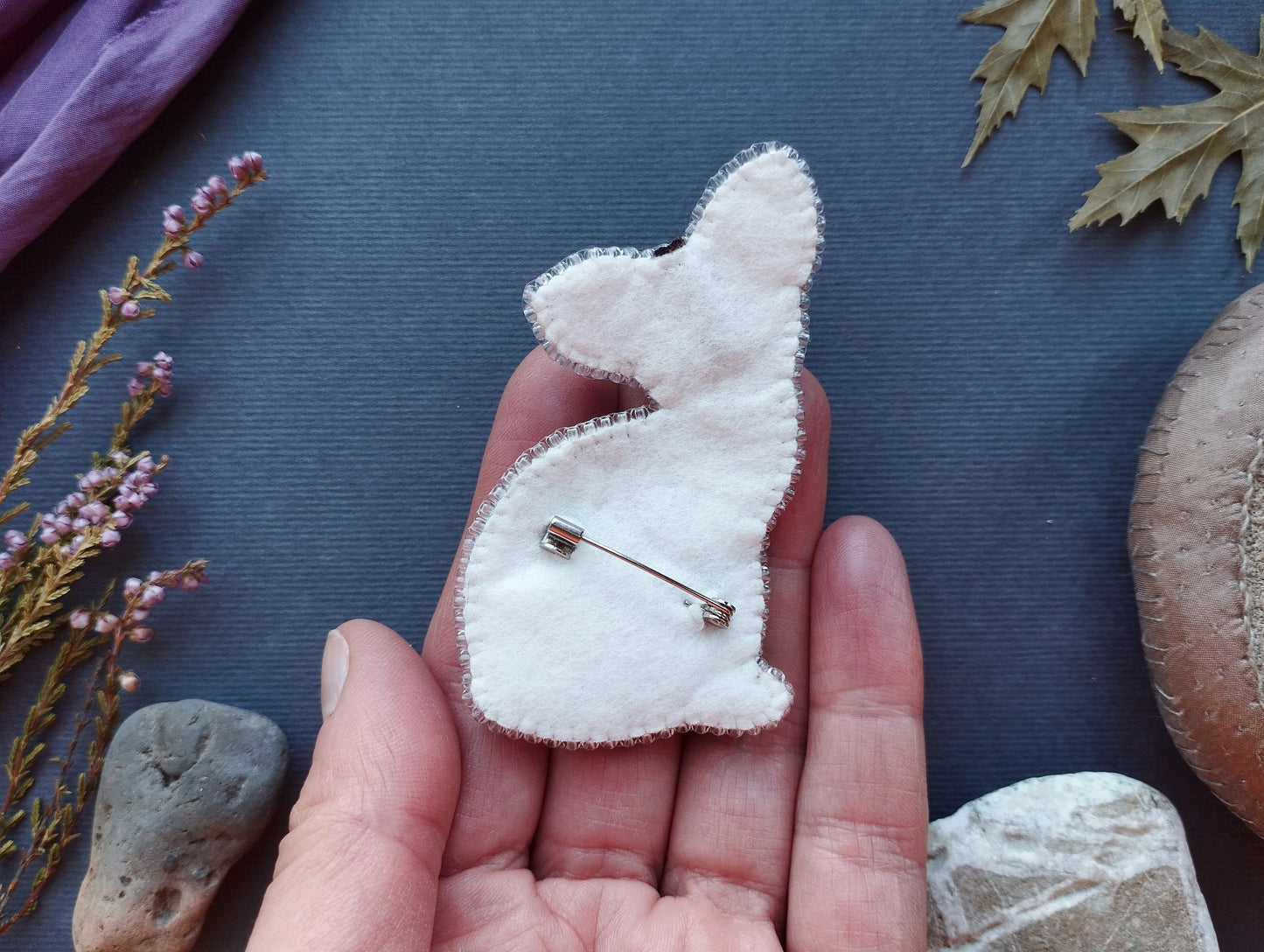 A hand holding a white felt rabbit pin against a dark background with various natural elements, such as leaves and rocks.
