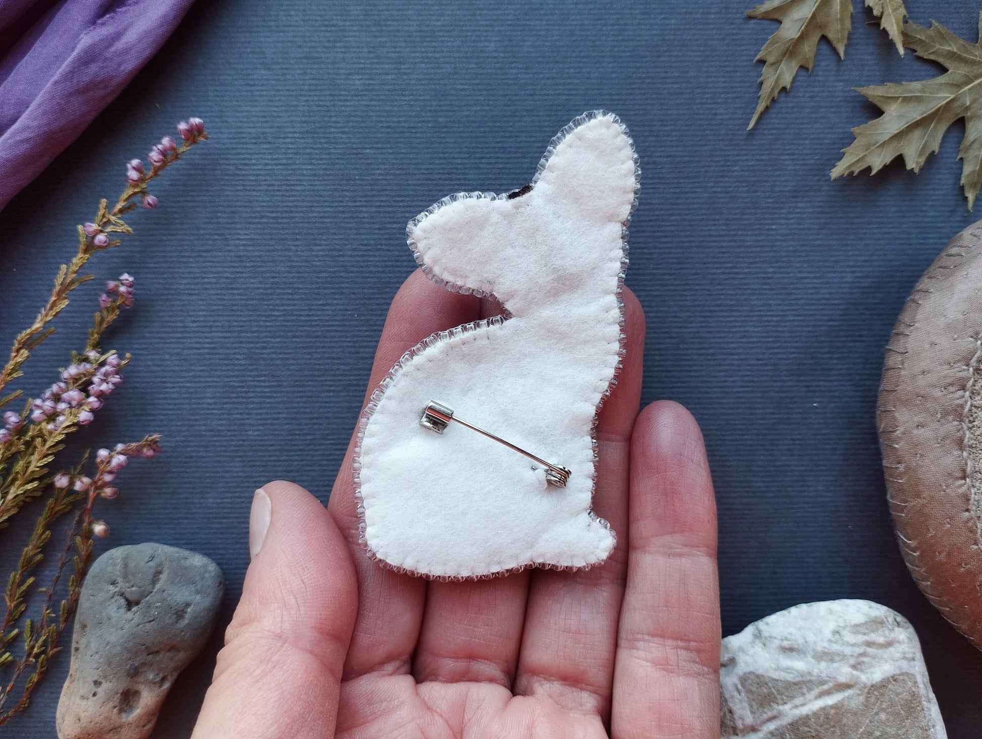 A hand holding a white felt rabbit pin against a dark background with various natural elements, such as leaves and rocks.