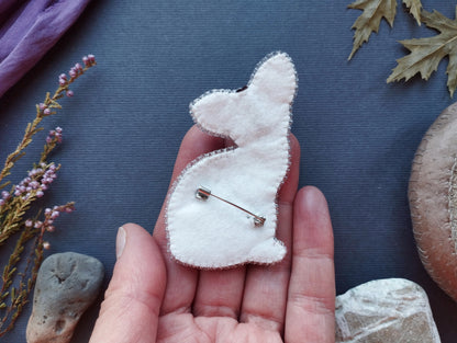 A hand holding a white felt rabbit pin against a dark background with various natural elements, such as leaves and rocks.