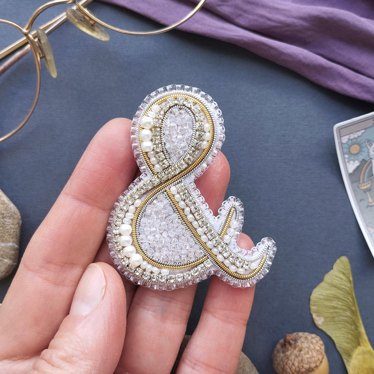 A hand holding a decorative beaded letter '&' on a dark background, with various other items such as glasses, a card, and a leaf visible in the background.